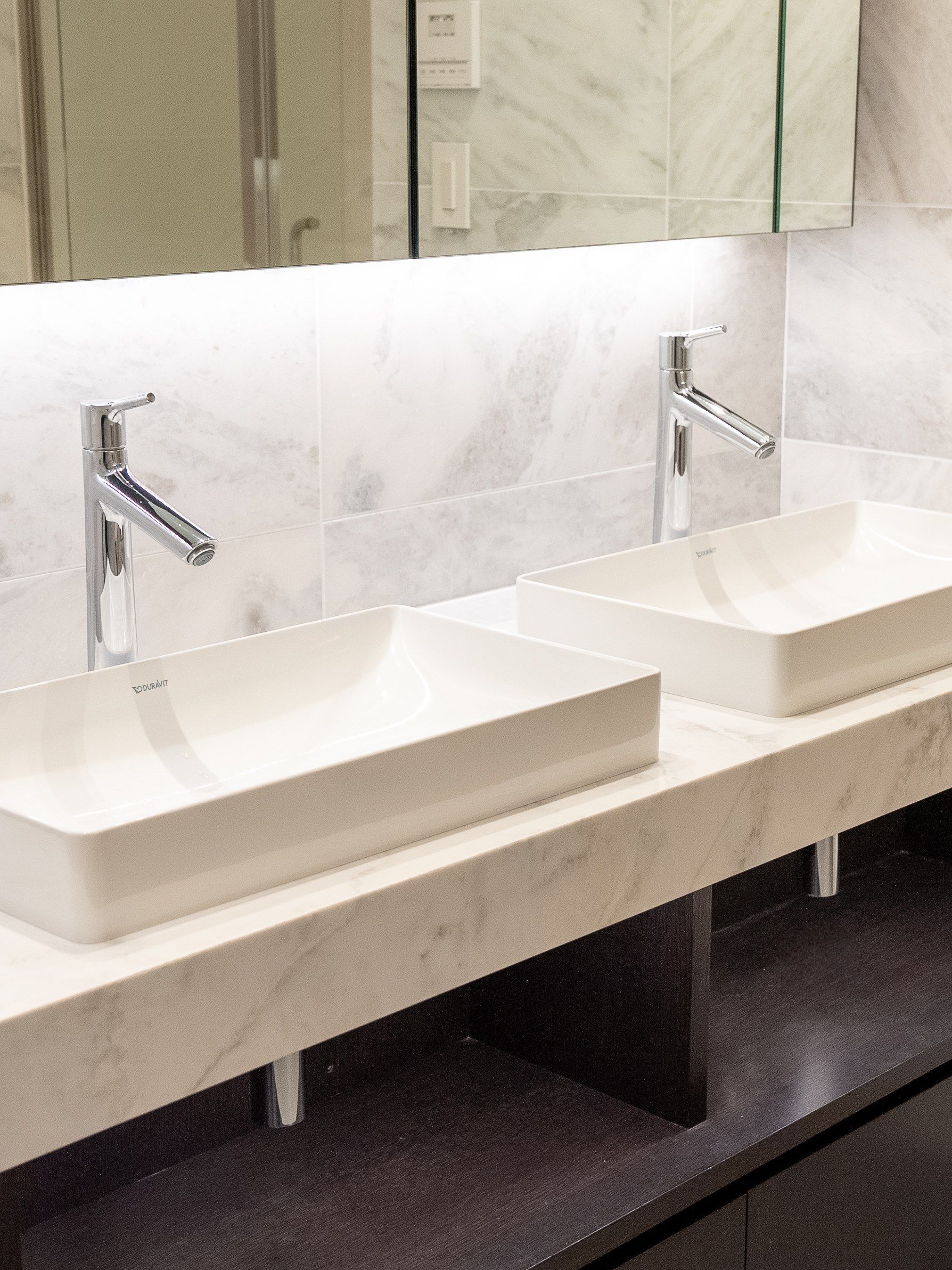 Two white basins on a marble counter, lit from behind the mirror.